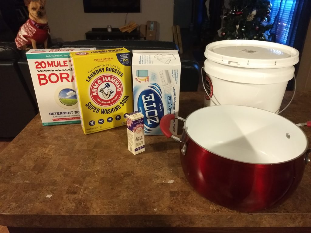 Materials for homemade laundry detergent assembled on a kitchen counter as a chihuahua wearing a red Christmas sweater looks on